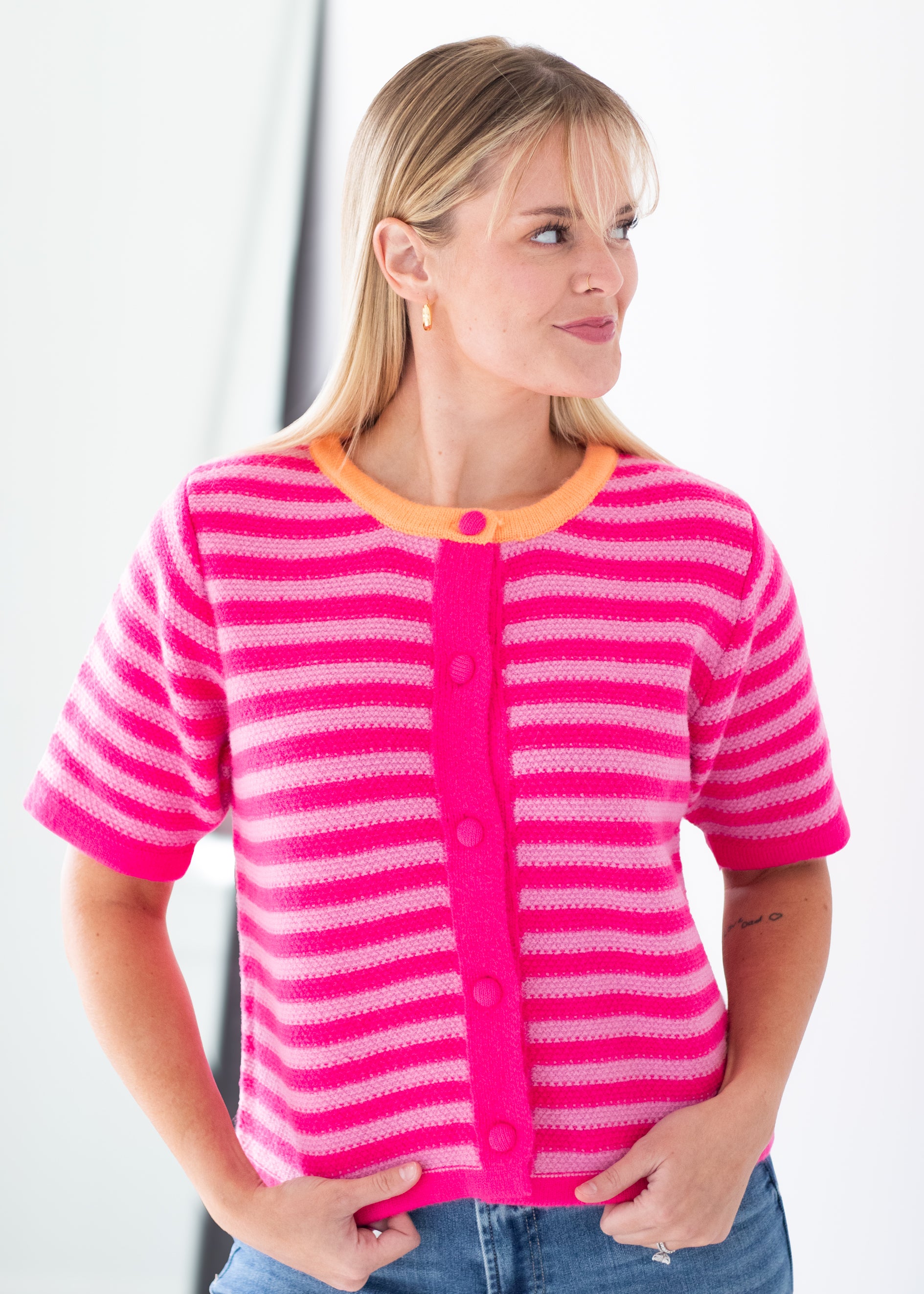 A woman wearing a short-sleeve knit cardigan with bright pink and white horizontal stripes, featuring a solid hot pink button placket and collar trim with a small orange accent.