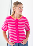 A woman wearing a short-sleeve knit cardigan with bright pink and white horizontal stripes, featuring a solid hot pink button placket and collar trim with a small orange accent.