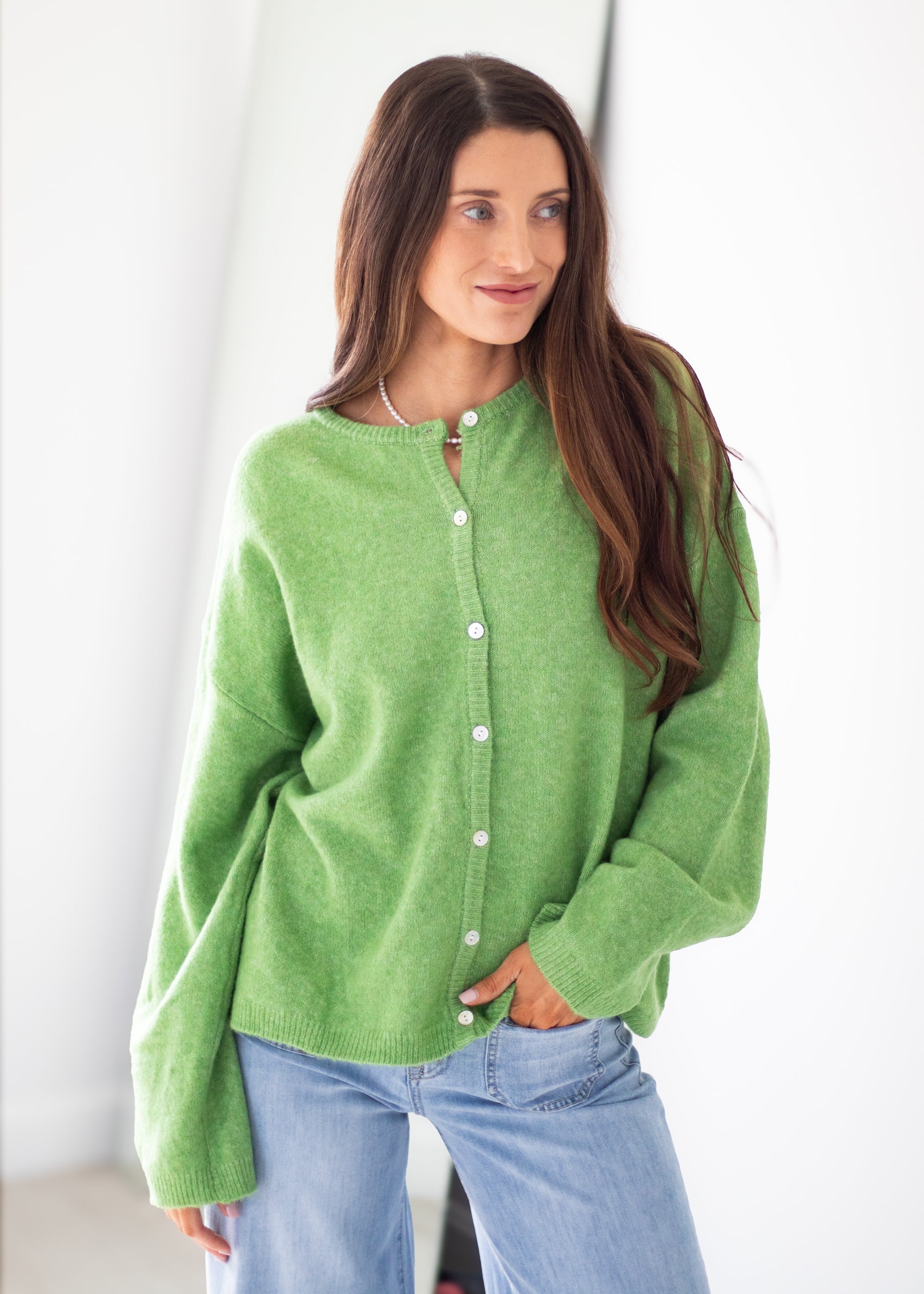 A woman wearing a fuzzy lime green button-up cardigan sweater and light blue jeans, standing against a bright white background.
