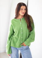 A woman wearing a fuzzy lime green button-up cardigan sweater and light blue jeans, standing against a bright white background.