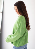 A woman with long dark brown hair is shown from the side, wearing a light green knit sweater and light blue jeans against a bright white background.