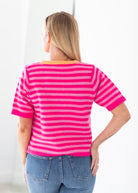 A woman viewed from behind wearing a bright pink and magenta striped short-sleeve knit top with an orange collar, paired with blue jeans, against a clean white background.