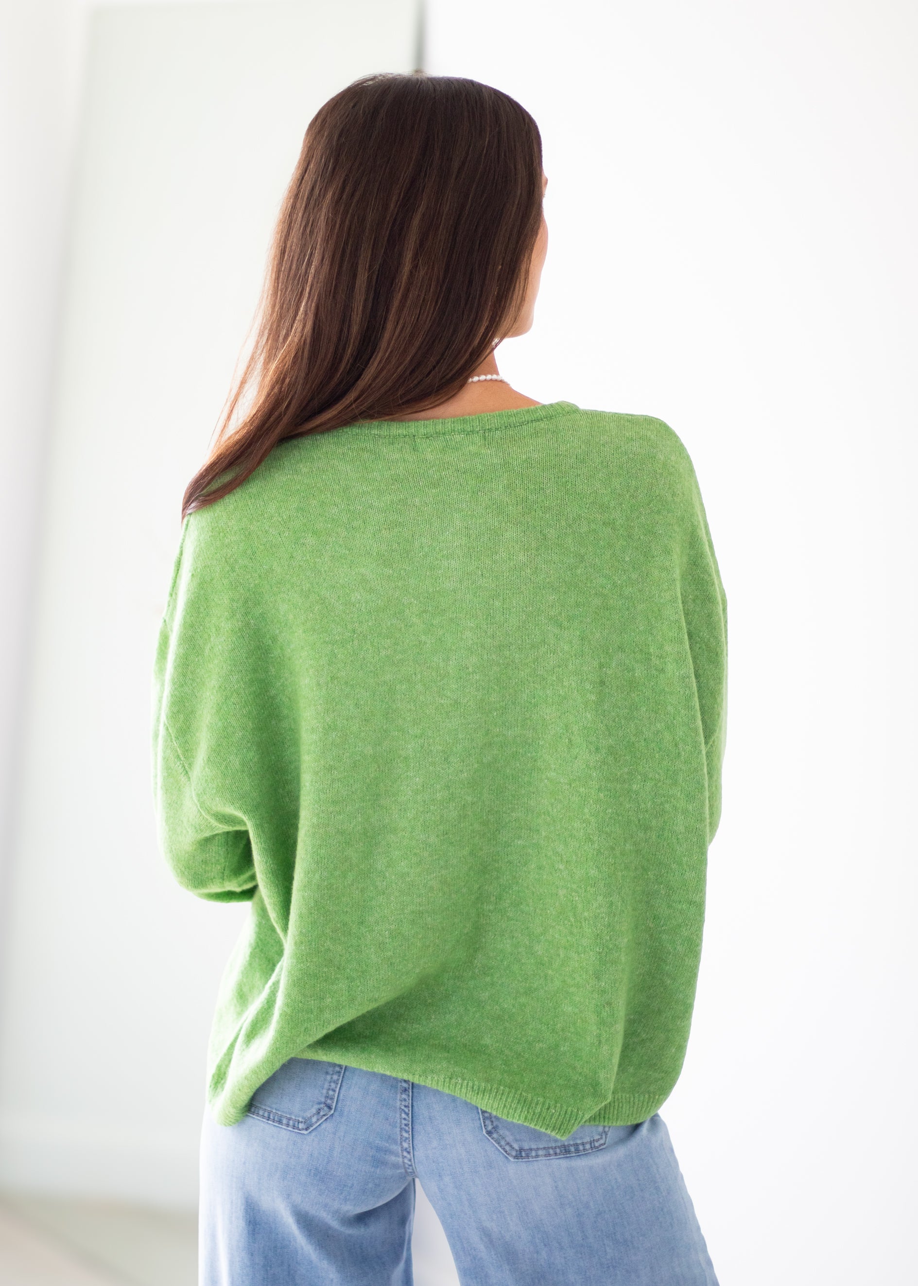 A woman viewed from behind wearing a bright green knit sweater and light blue jeans against a clean white background.