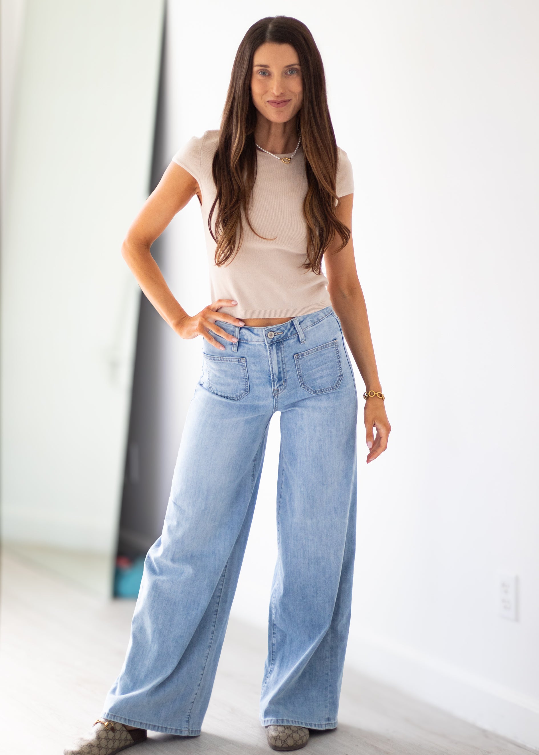 A woman modeling a light beige short-sleeve crop top and light-wash wide-leg blue jeans, standing in a bright indoor setting with a white wall and light floor.