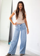 A woman modeling a light beige short-sleeve crop top and light-wash wide-leg blue jeans, standing in a bright indoor setting with a white wall and light floor.