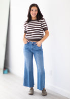 A woman modeling a brown and white horizontally striped short-sleeve knit top with blue wide-leg jeans and brownish-gray slip-on shoes, standing in a bright indoor room with a white wall.