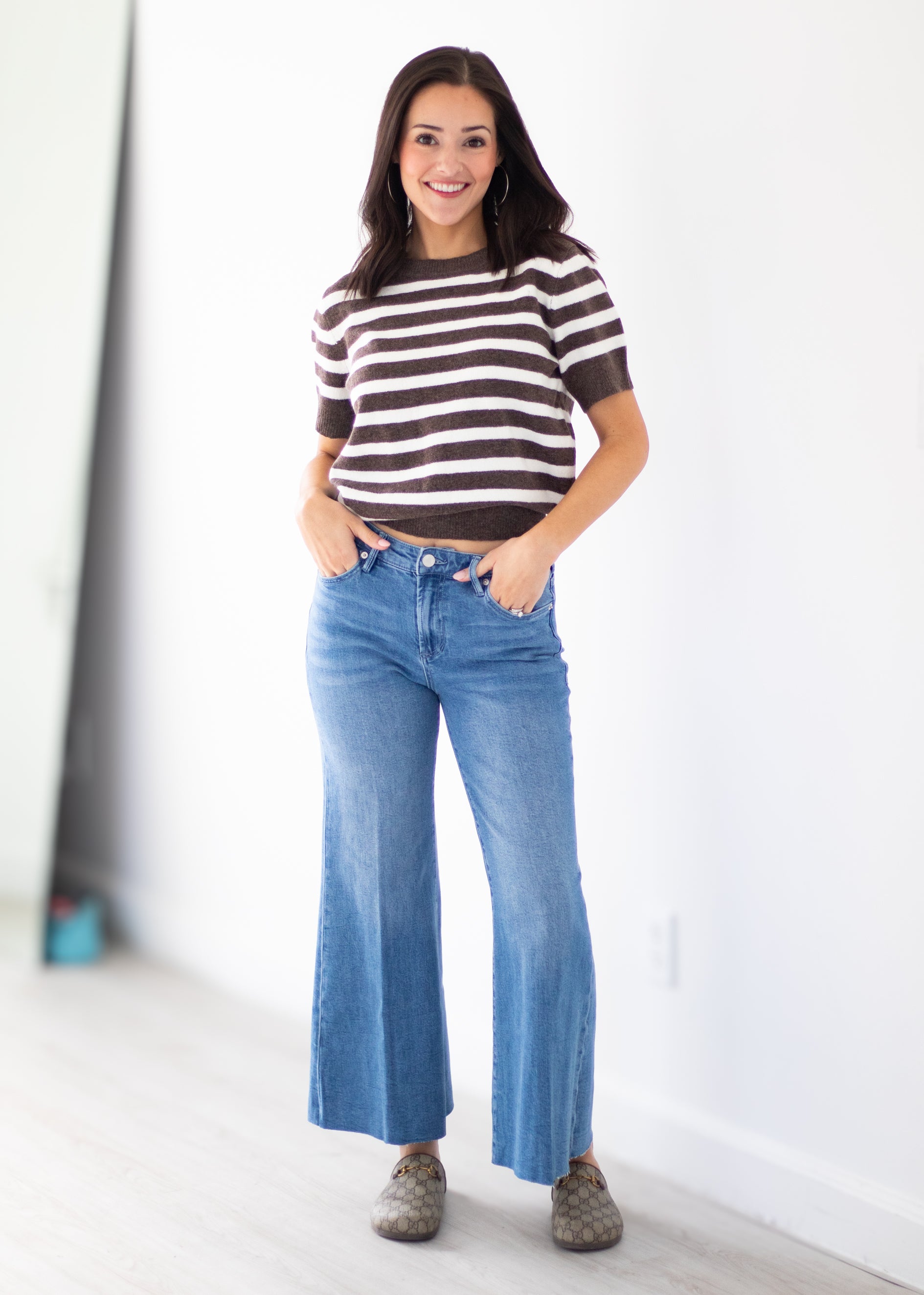 A woman modeling a brown and white horizontally striped short-sleeve knit top with blue wide-leg jeans and brownish-gray slip-on shoes, standing in a bright indoor room with a white wall.