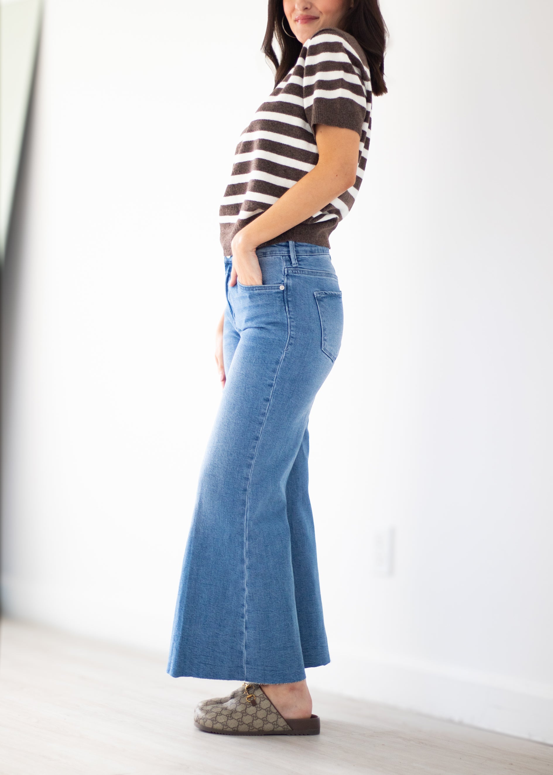 A woman wearing a brown and white striped short-sleeve top, a high-waisted blue flare jeans, and brown patterned slip-on shoes, standing in a bright room with white walls and light wood flooring.