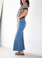 A woman wearing a brown and white striped short-sleeve top, a high-waisted blue flare jeans, and brown patterned slip-on shoes, standing in a bright room with white walls and light wood flooring.