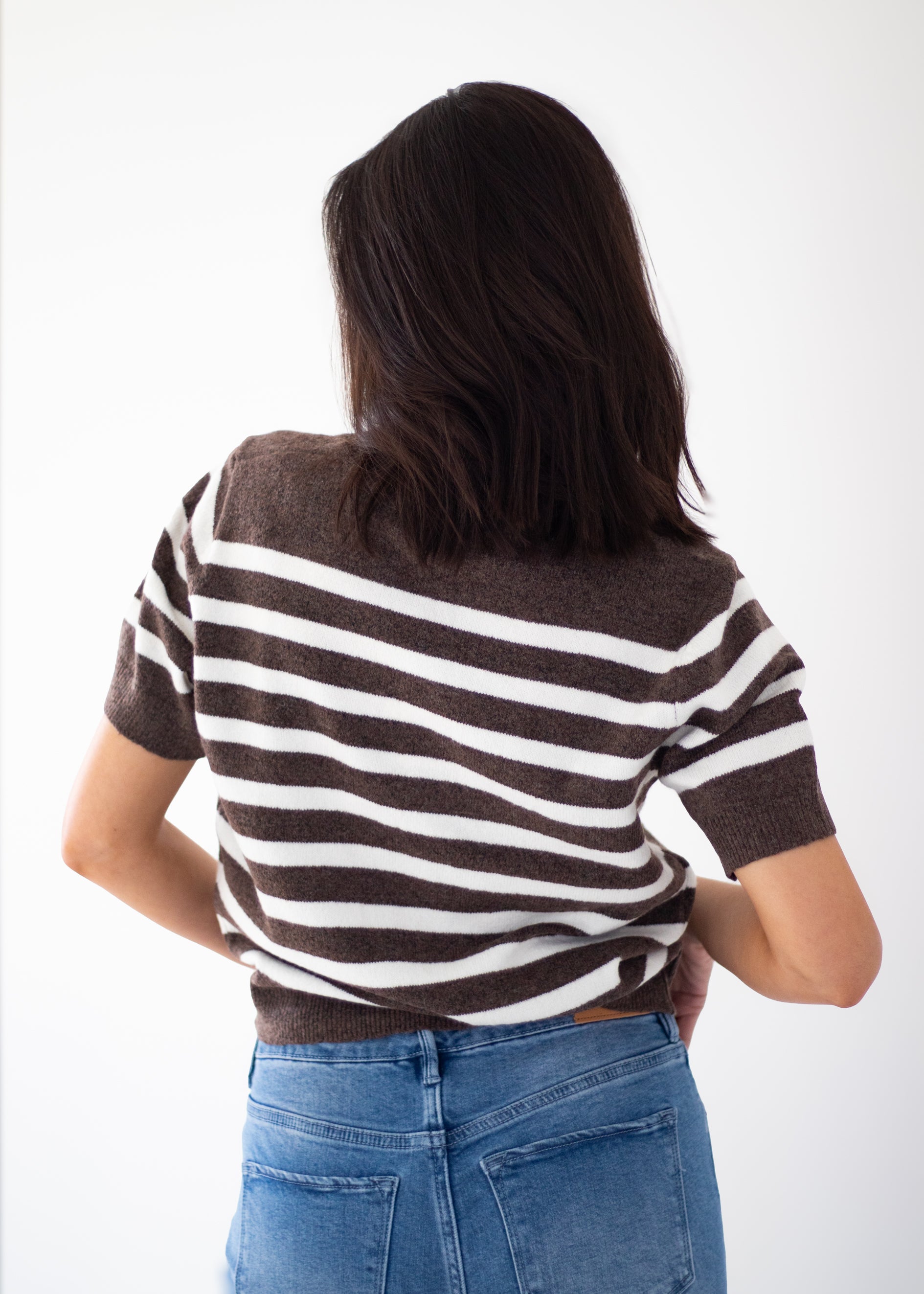 A woman viewed from behind wearing a brown and white horizontally striped knit top with blue jeans, set against a plain white studio background.