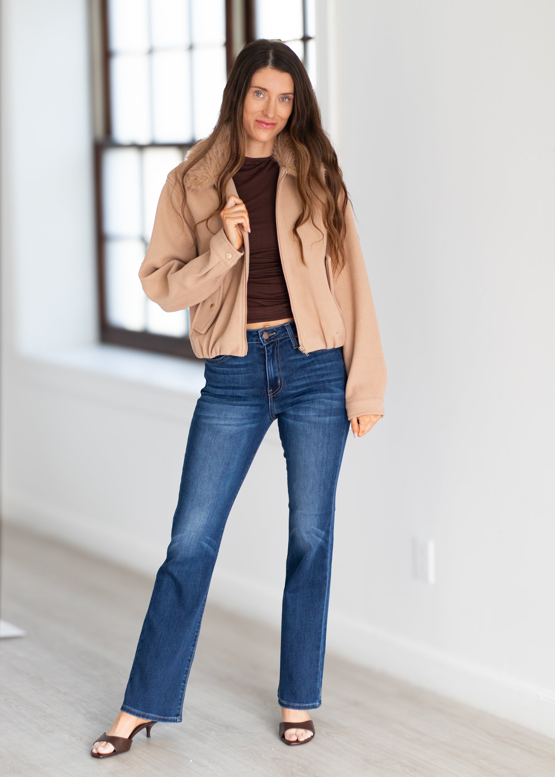 A woman stands in a bright room wearing a tan jacket, dark brown ribbed top, blue flare jeans, and brown heeled sandals.
