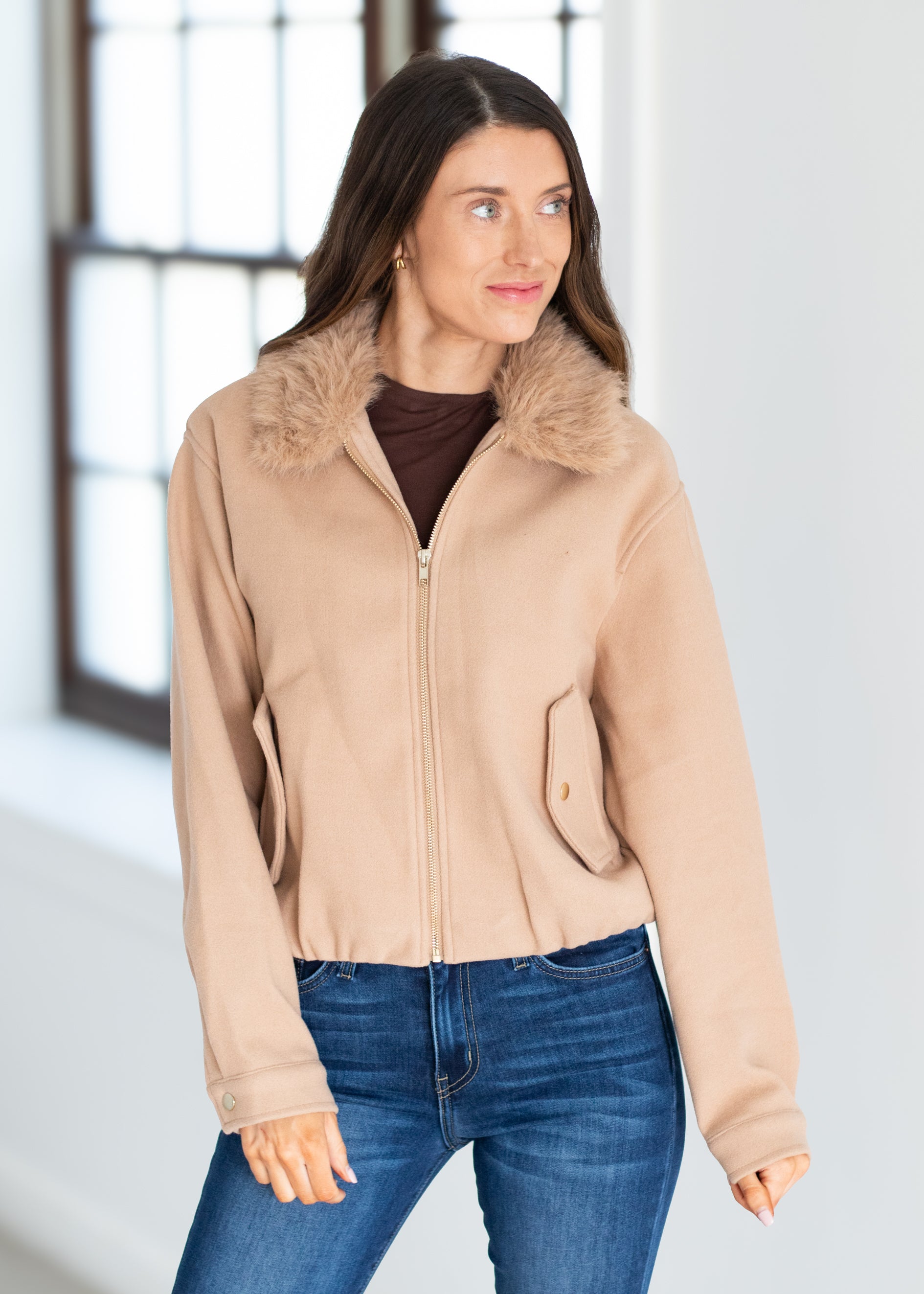 A woman wearing a beige cropped jacket with a fuzzy faux-fur collar, paired with a dark brown top and blue jeans, standing in a brightly lit room with a window in the background.