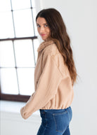 A woman with long brown hair is shown from the back and side, wearing a light beige cropped sweatshirt with a fuzzy collar and blue jeans, standing in a bright room with a large window.