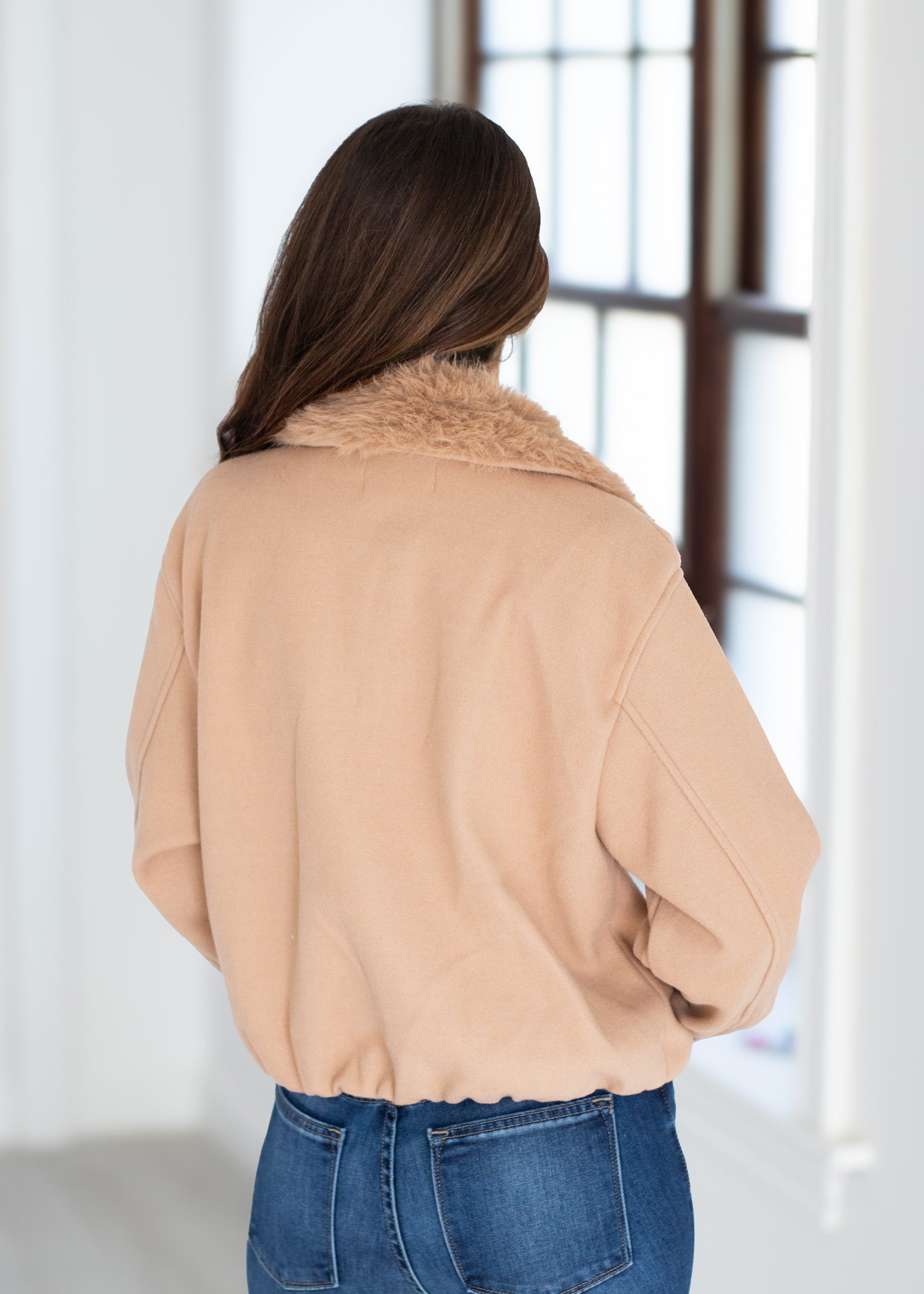 A woman viewed from behind wearing a light tan cropped jacket with a fuzzy collar and blue jeans, standing in a bright indoor setting near a window.