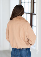A woman viewed from behind wearing a light tan cropped jacket with a fuzzy collar and blue jeans, standing in a bright indoor setting near a window.