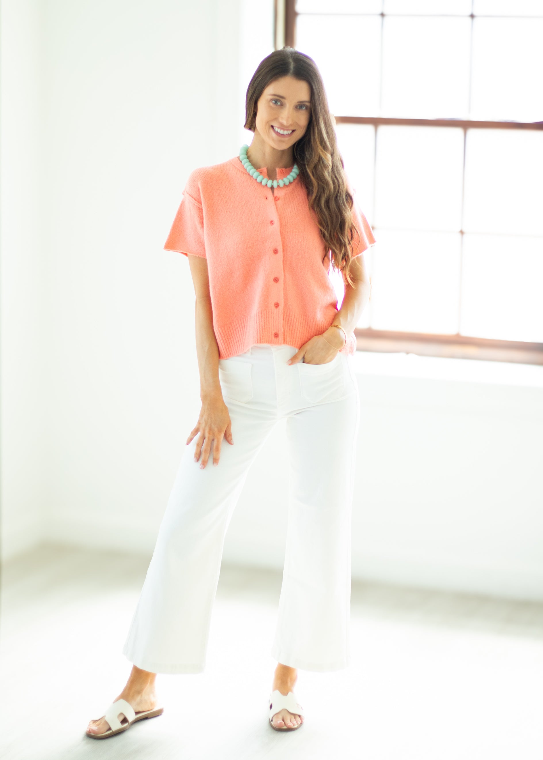 Woman wearing a coral blouse and white pants in a bright room.