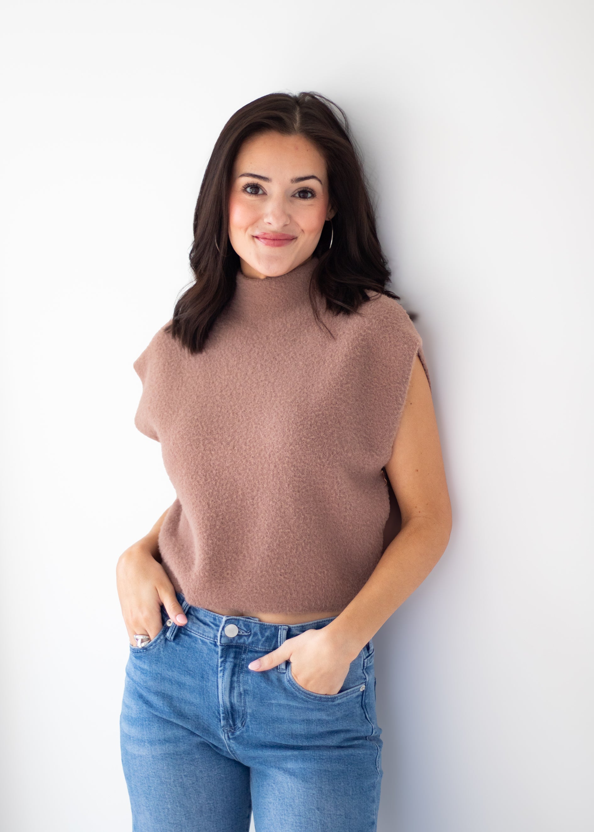 A woman with dark hair wearing a sleeveless, fuzzy taupe-brown mock neck top and blue jeans, standing against a plain white wall.
