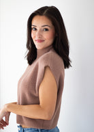 A woman with dark brown hair is shown from a side-back angle wearing a sleeveless, high-neck cropped sweater in a dusty mauve-brown color, paired with blue jeans, against a plain white studio background.