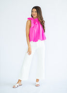 Woman wearing a bright pink blouse with a bow and white pants on a light gray background