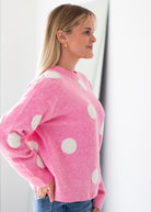 A woman in profile wearing a pink knit sweater with large white polka dots, paired with blue jeans, standing against a bright white wall.