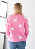 A woman viewed from behind wearing a pink knit sweater with large white polka dots and blue jeans, standing in a bright indoor setting.