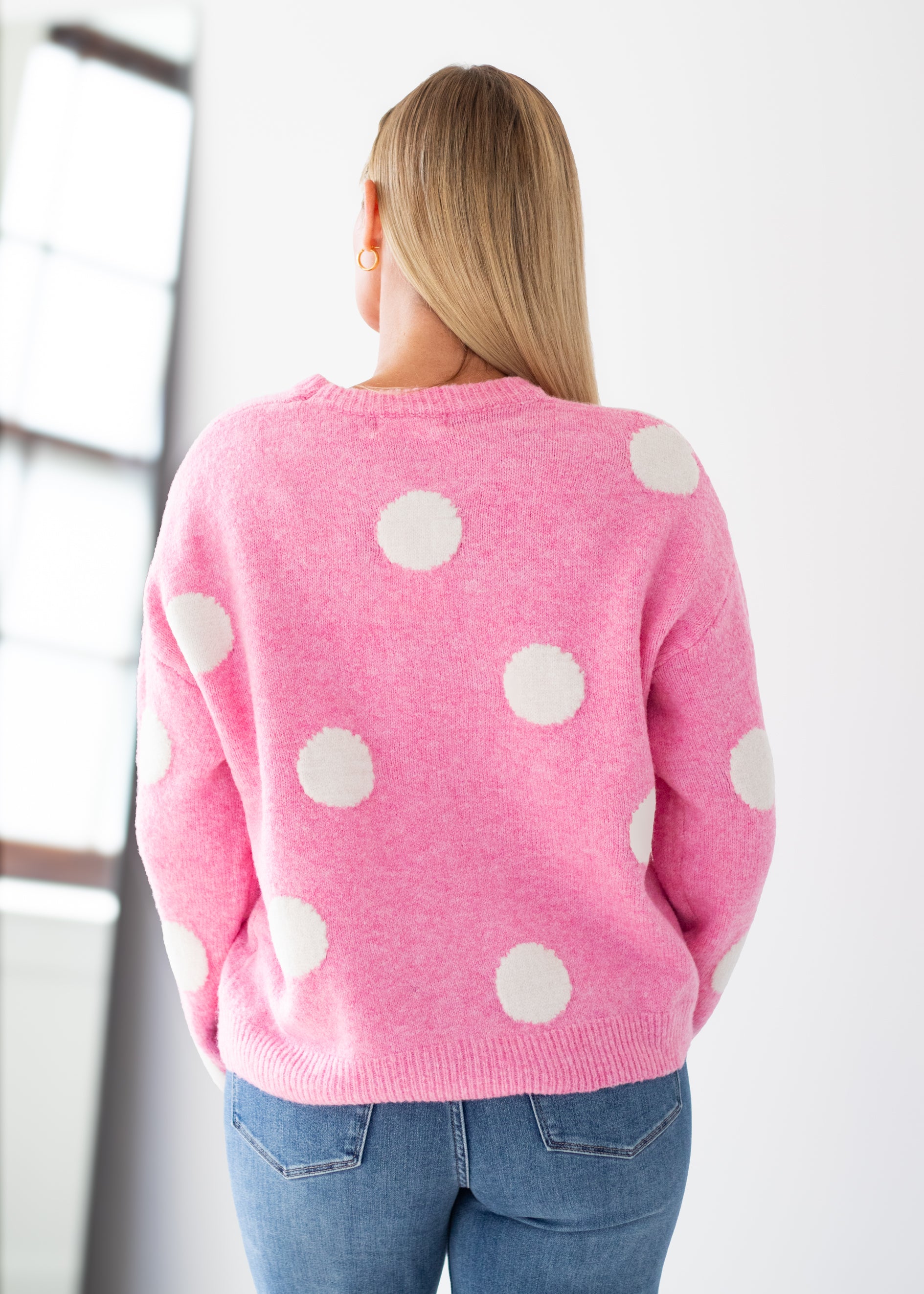 A woman viewed from behind wearing a pink knit sweater with large white polka dots and blue jeans, standing in a bright indoor setting.