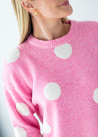 A close-up of a woman wearing a pink knit sweater with large white polka dots, shown from the shoulders to the chest against a light neutral background.