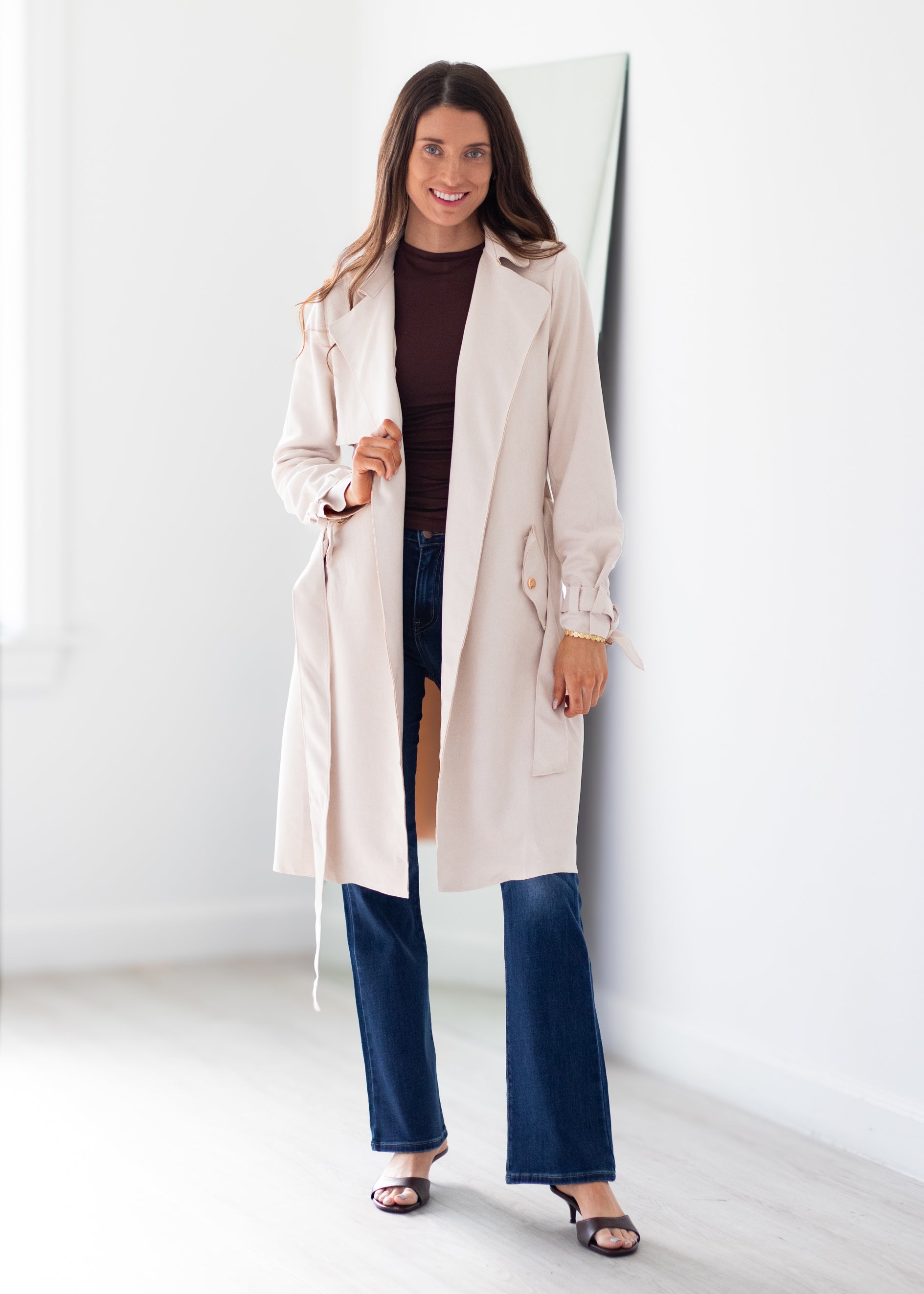 A woman modeling a light beige trench coat over a dark brown top and blue jeans, standing in a bright, minimalist room with a white wall and floor.