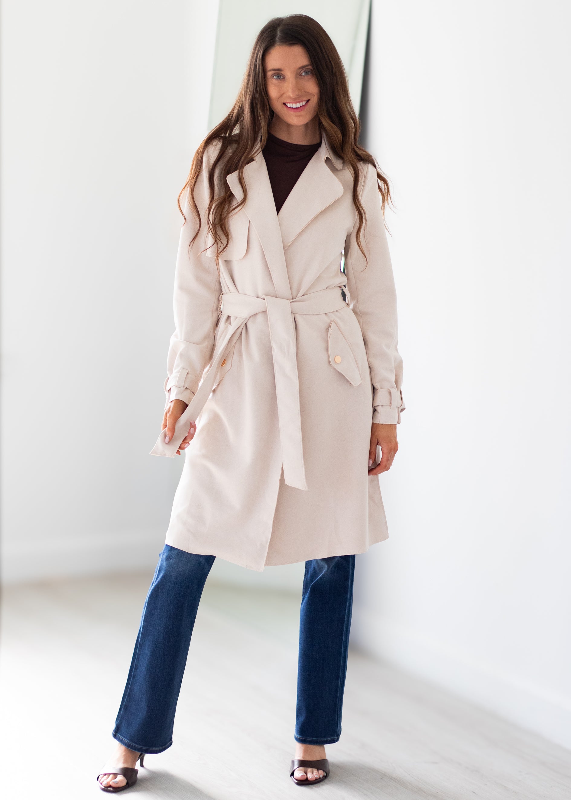 A woman modeling a light beige trench coat with a tie waist belt, worn over a dark brown top and blue jeans, standing in a bright, minimalist white room.