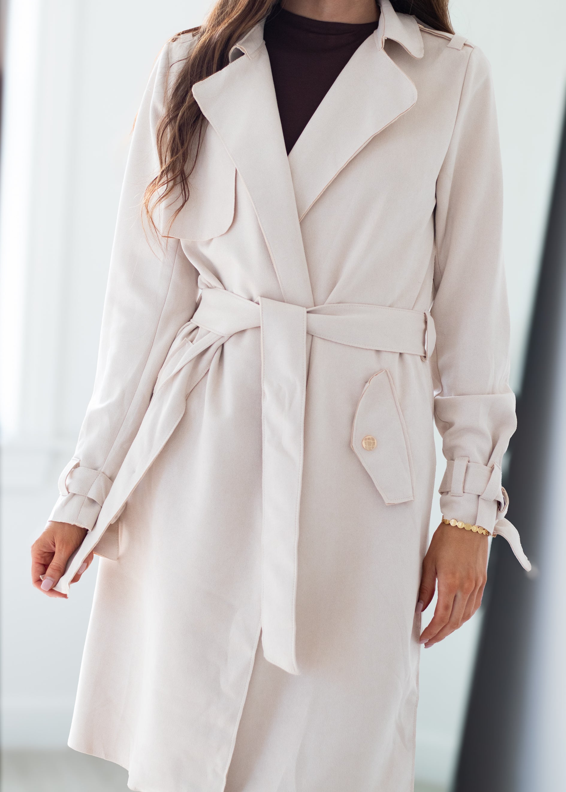 A woman wearing a light beige trench coat with a matching waist tie belt, styled over a dark brown top. The photo is cropped from the chin down, set against a bright, softly blurred indoor background.