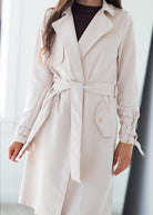 A woman wearing a light beige trench coat with a matching waist tie belt, styled over a dark brown top. The photo is cropped from the chin down, set against a bright, softly blurred indoor background.