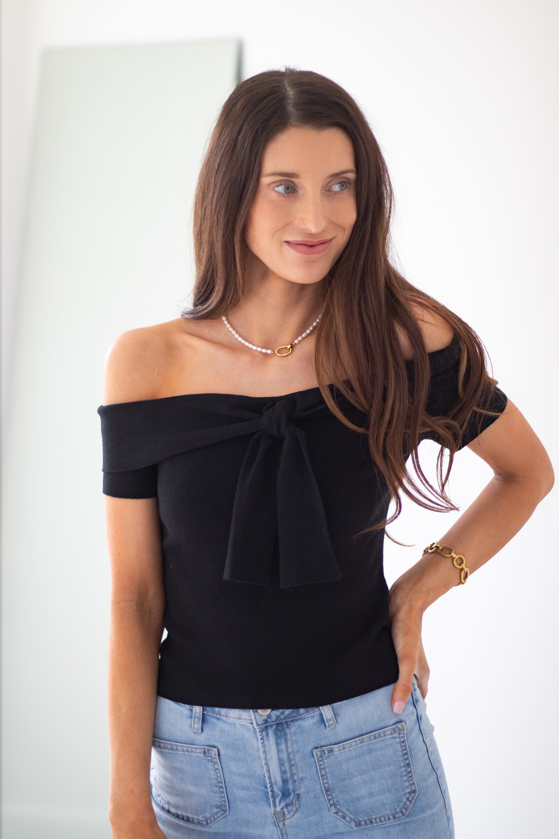 A woman modeling a black off-the-shoulder top with a large bow detail at the neckline, paired with light blue jeans, against a plain white background.