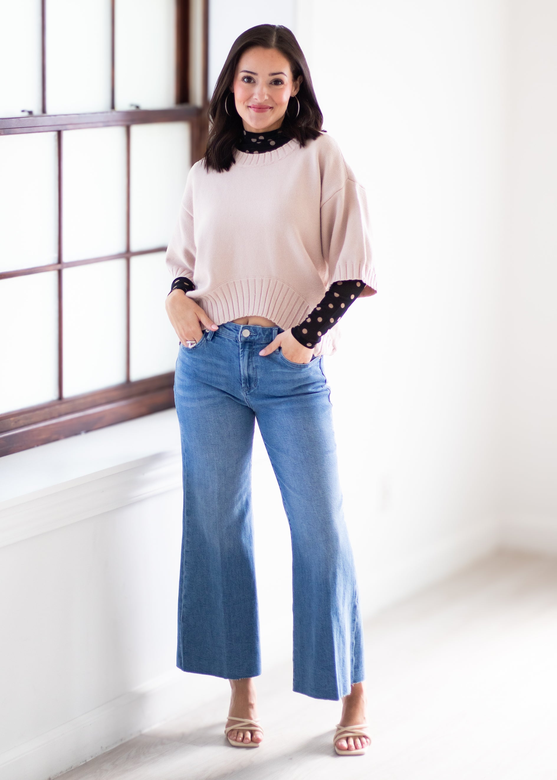 A woman modeling a light beige knit sweater with black polka-dot and lace-trimmed sleeves, paired with blue wide-leg jeans and beige sandals, standing in a bright room with a window.