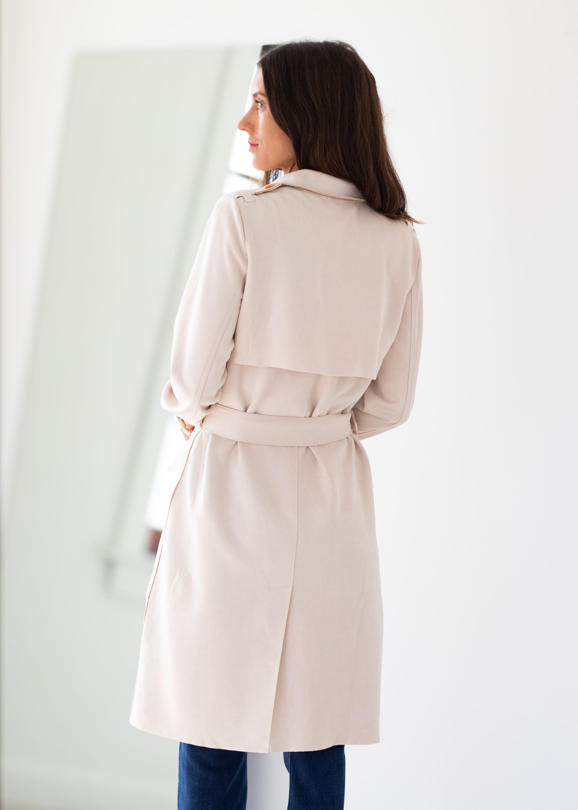 A woman wearing a light beige trench coat with a matching belt, shown from the back in a bright, minimalist indoor setting.