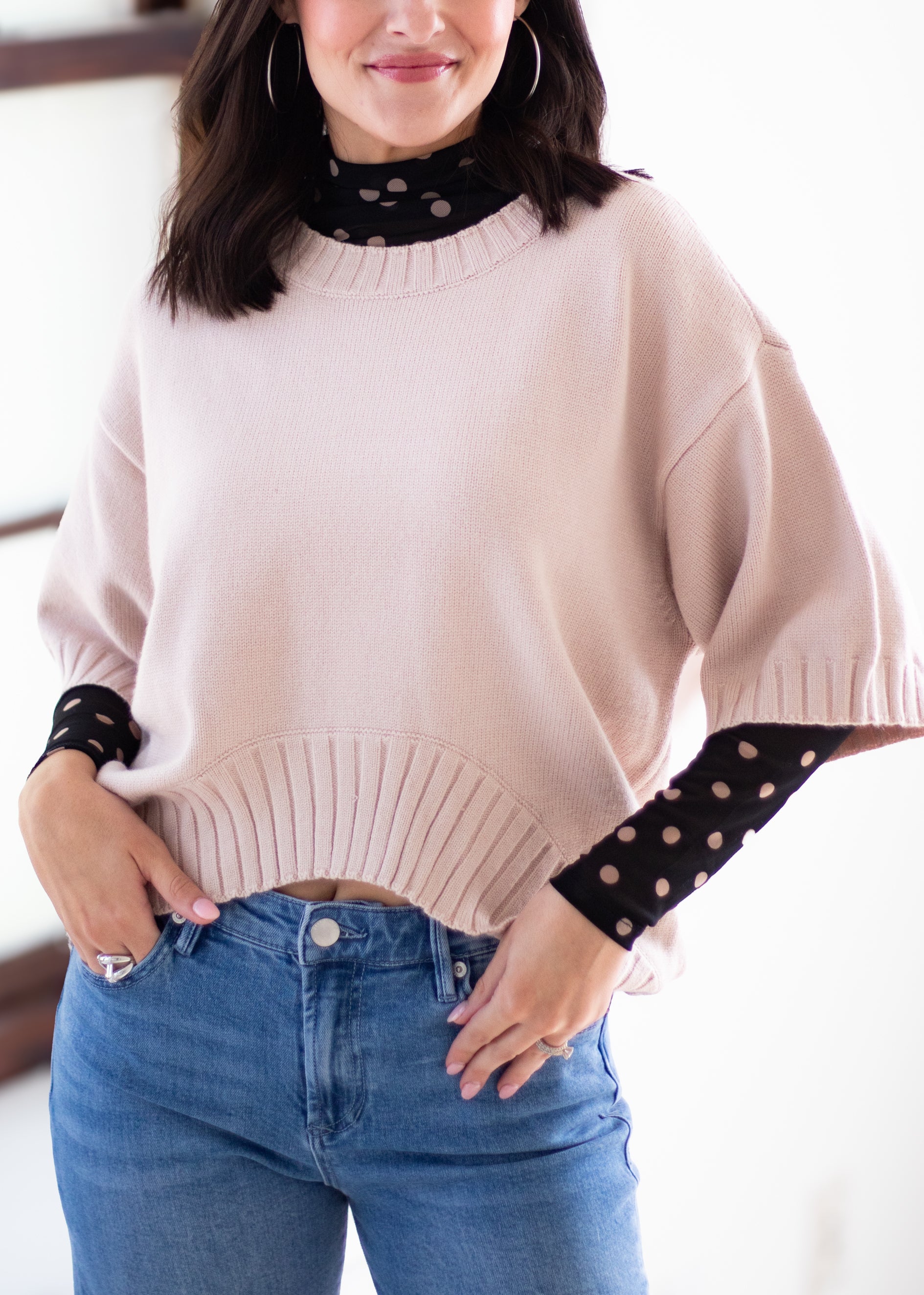 A woman wearing a light blush pink knit sweater with wide short sleeves and a ribbed hem, layered over a black sheer long-sleeve top with beige polka dots, paired with blue jeans.