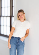 A woman wearing a white short-sleeve button-front knit top and blue jeans, standing indoors against a white wall near a window.