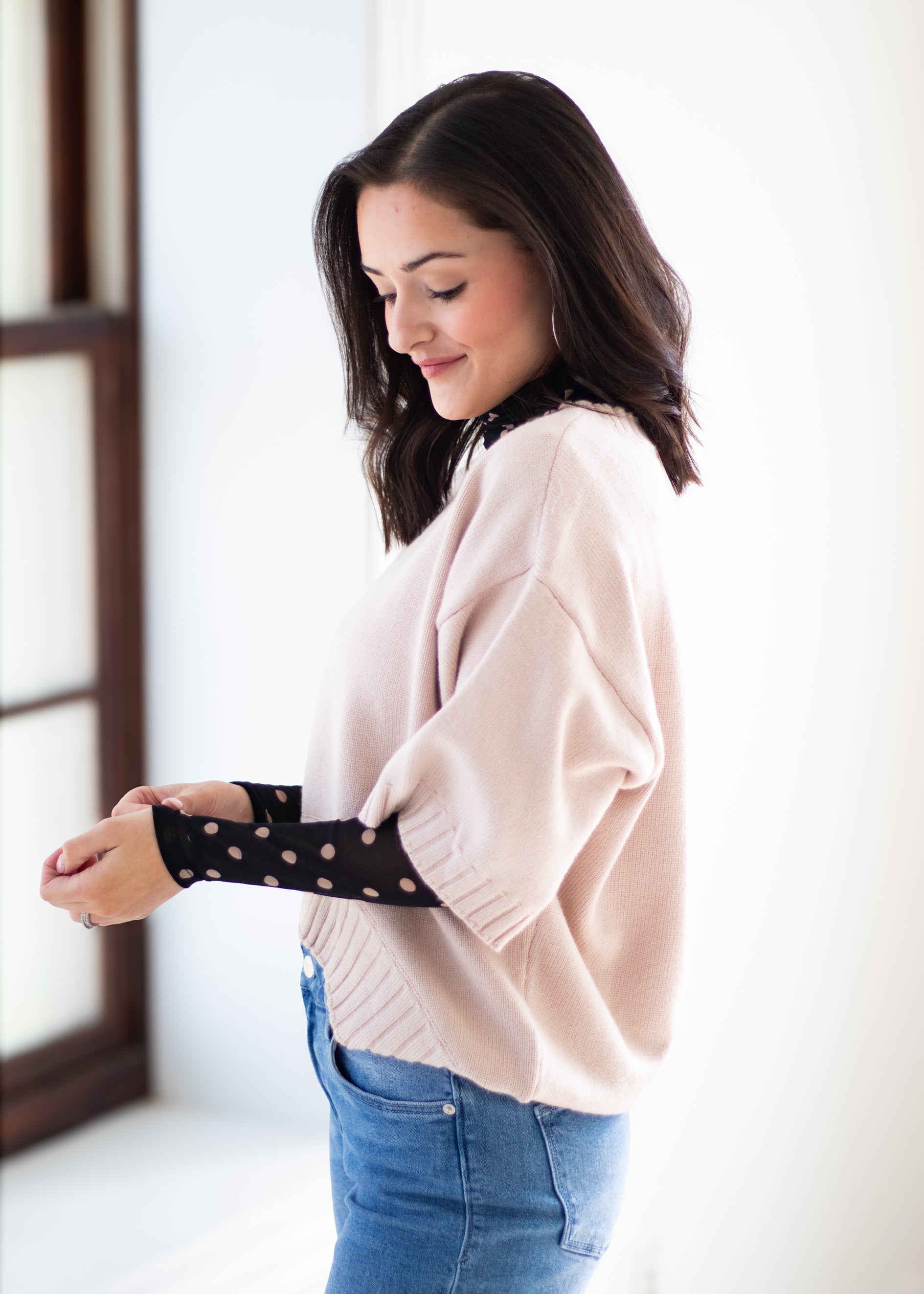 A woman wearing a light pink knit sweater and blue jeans, adjusting a black long-sleeve shirt with pink polka dots. The photo is taken in a brightly lit indoor setting.
