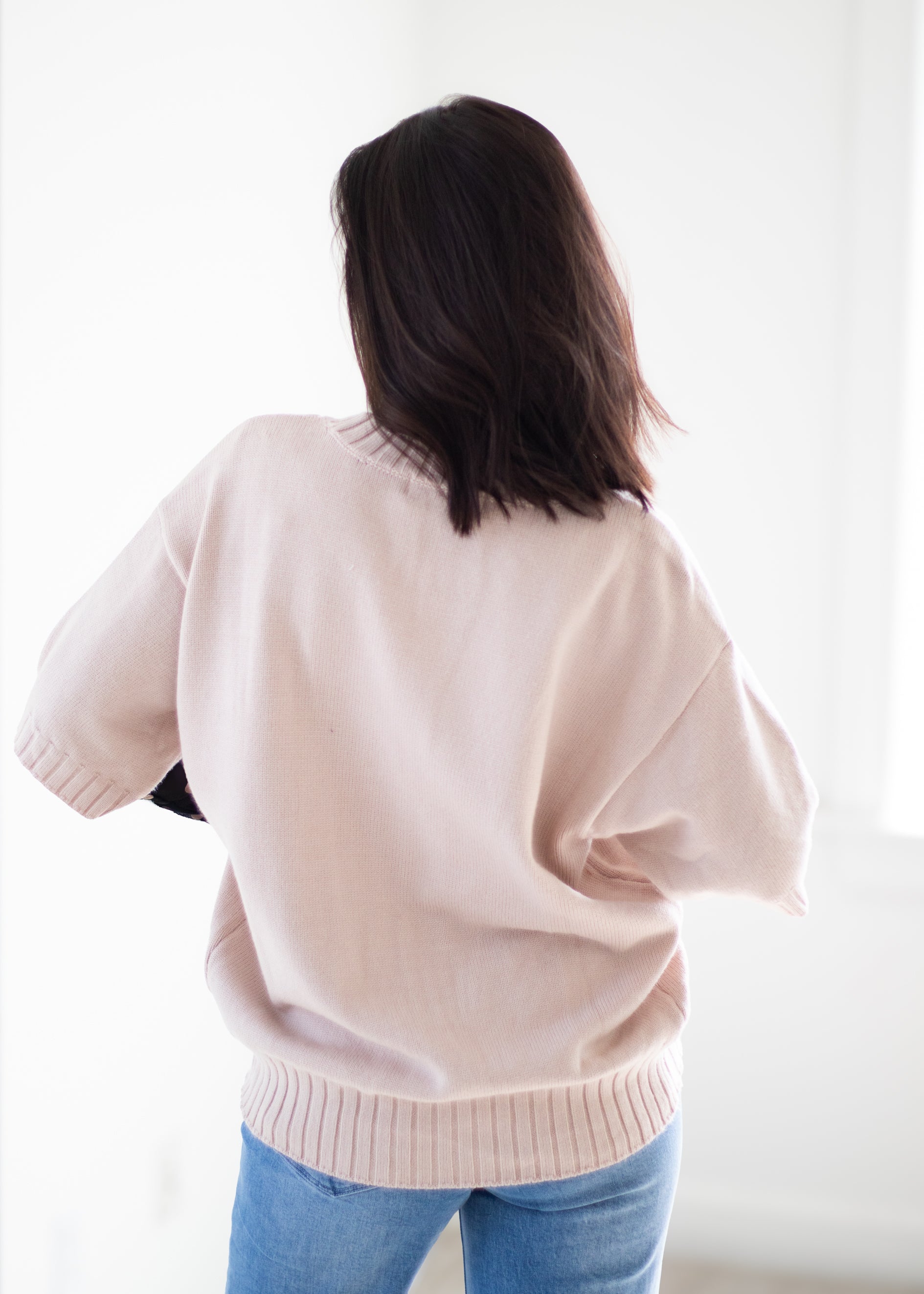 A woman viewed from behind wearing a light blush pink knit sweater and blue jeans against a bright white background.