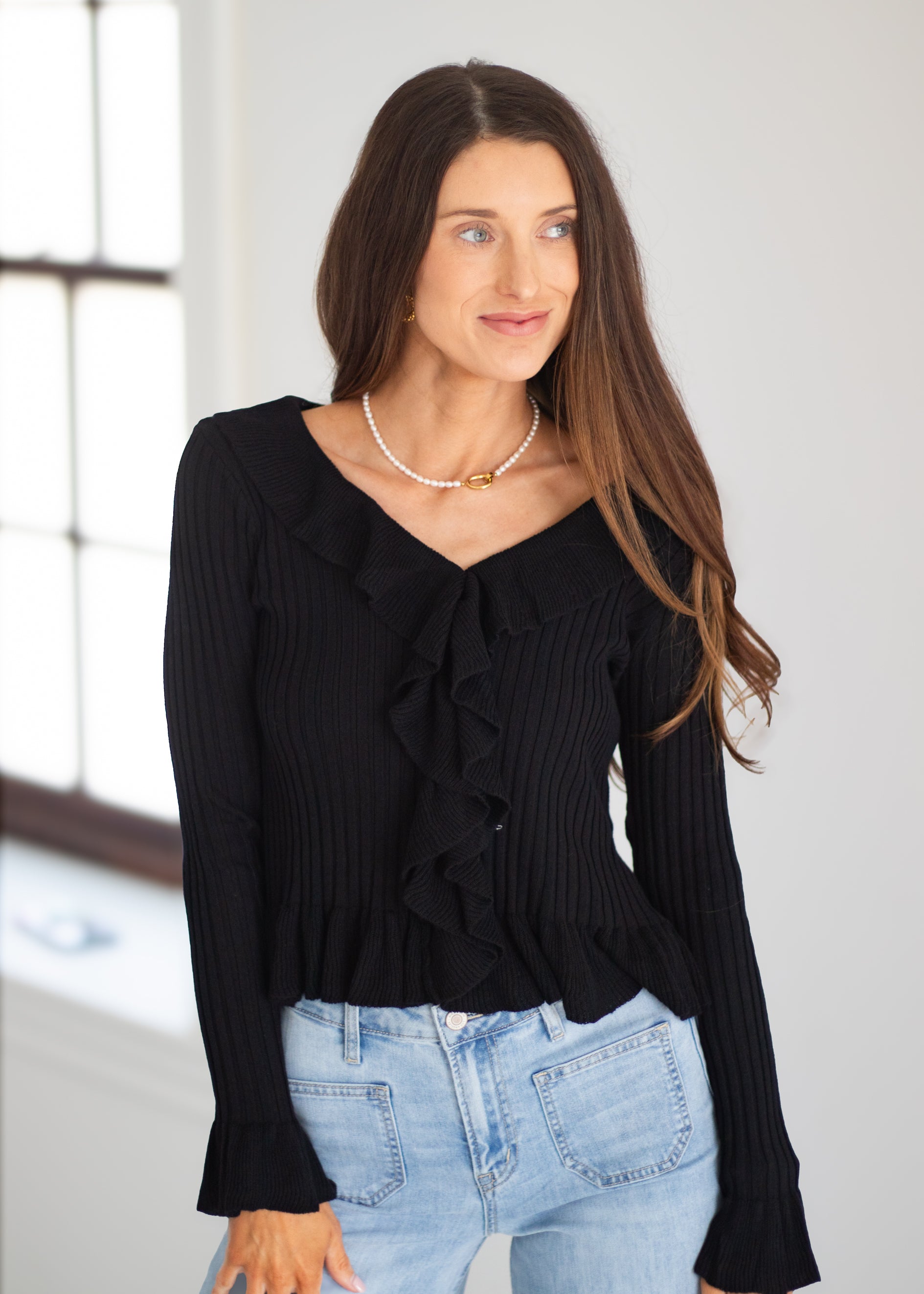 A woman modeling a black long-sleeve ribbed top with a ruffled V-neckline, paired with light blue jeans, in a bright indoor setting.