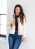 A woman modeling a cream-colored jacket with brown and black gingham trim, worn over a dark brown top and blue jeans, standing in a bright indoor setting.