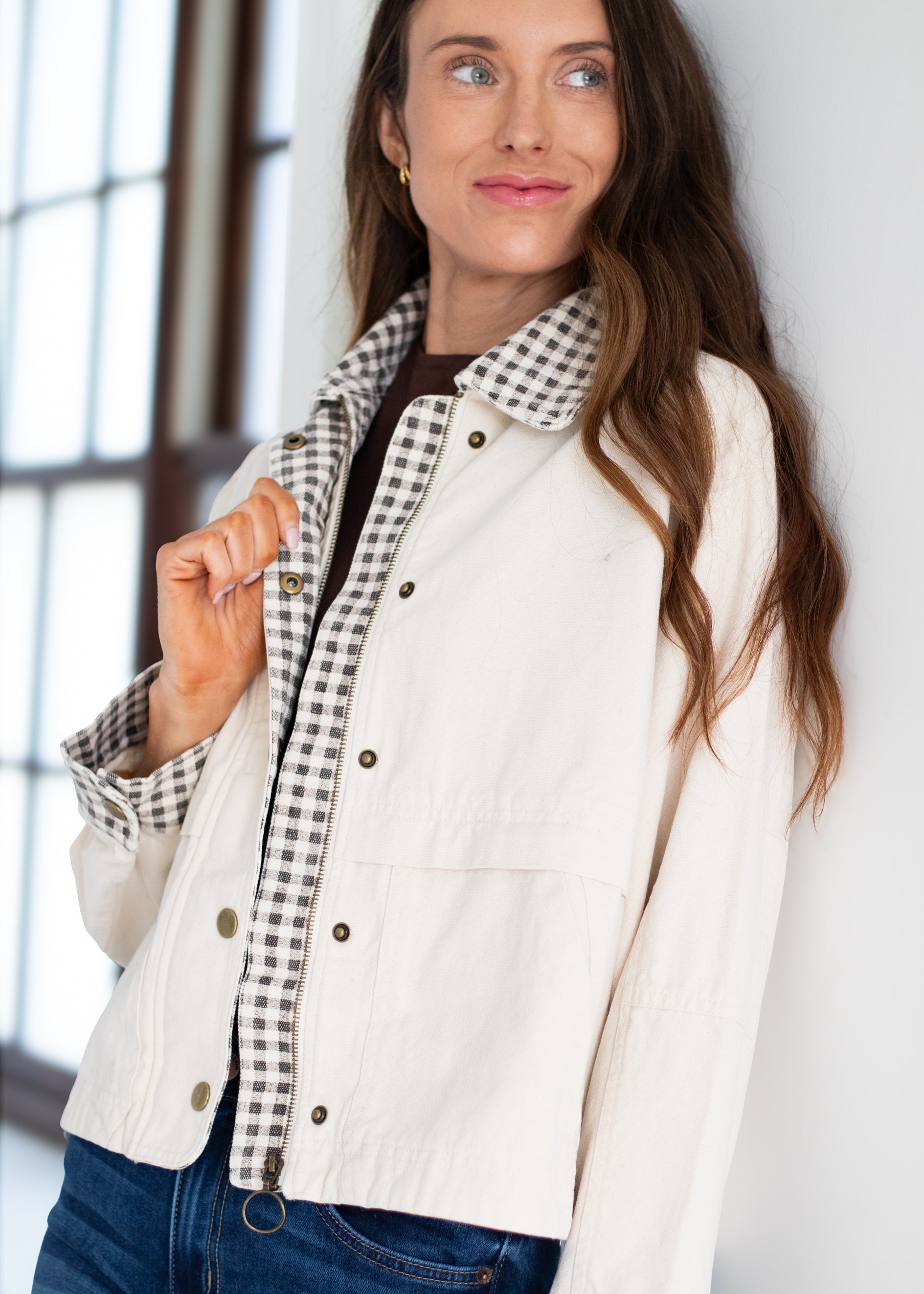A woman wearing a cream-colored jacket with a black and white gingham patterned lining, styled with a dark brown shirt and blue jeans.