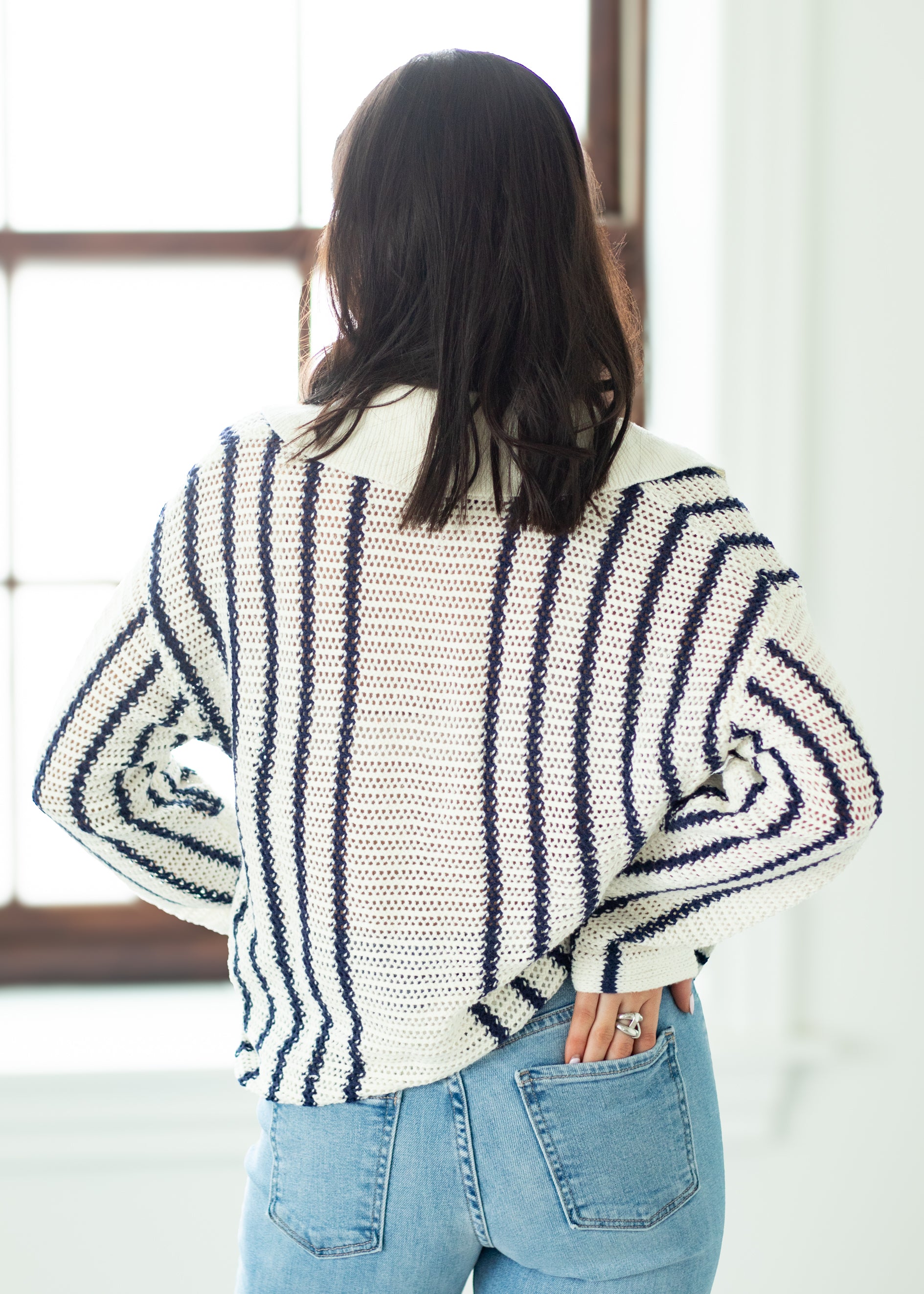 Person wearing a striped sweater and blue jeans in a room with a window.