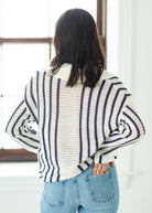Person wearing a striped sweater and blue jeans in a room with a window.