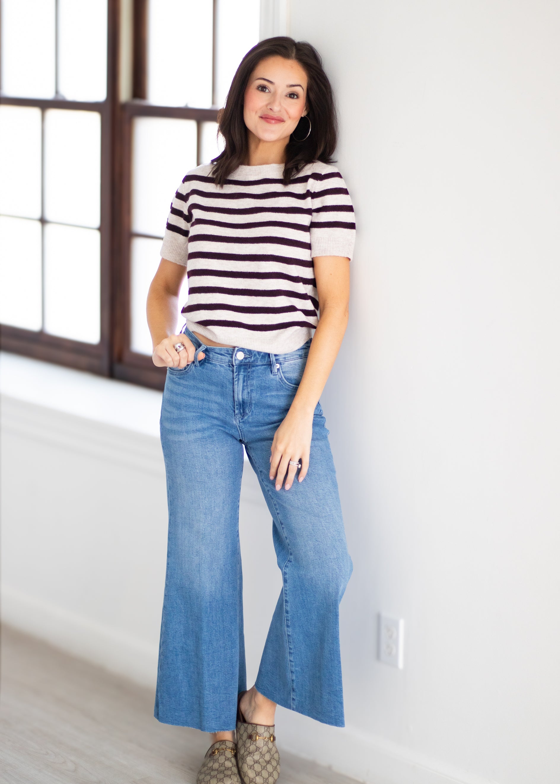 A woman stands indoors wearing a short-sleeve striped knit top with dark brown and off-white horizontal stripes, paired with light blue wide-leg jeans and brown patterned shoes.