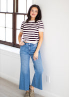 A woman stands indoors wearing a short-sleeve striped knit top with dark brown and off-white horizontal stripes, paired with light blue wide-leg jeans and brown patterned shoes.
