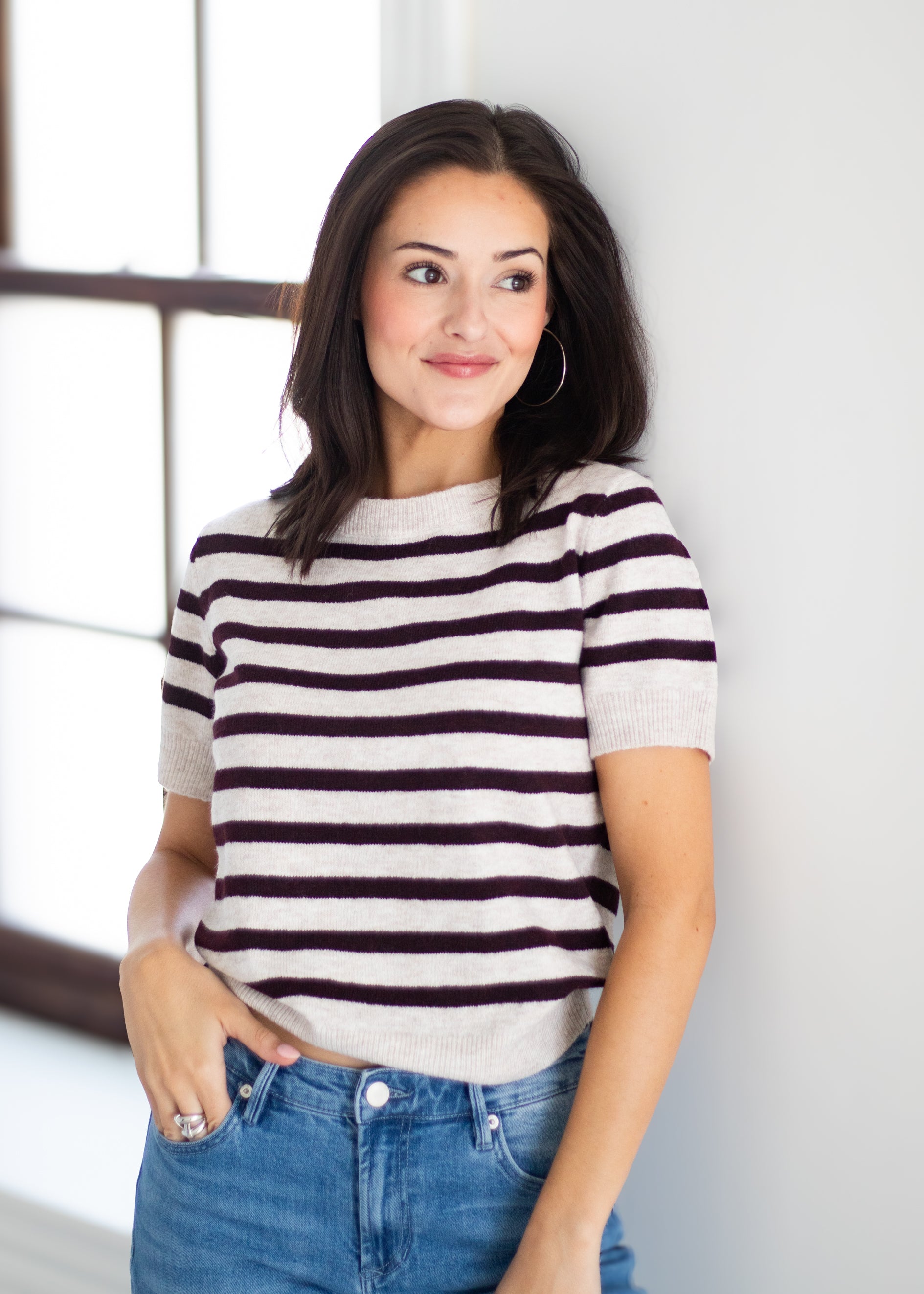 A woman wearing a short-sleeve knit top with horizontal dark brown and cream stripes, paired with blue jeans, standing against a white wall.