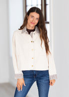 A woman modeling a cream-colored jacket with brown and white gingham patterned collar and cuffs, paired with blue jeans.