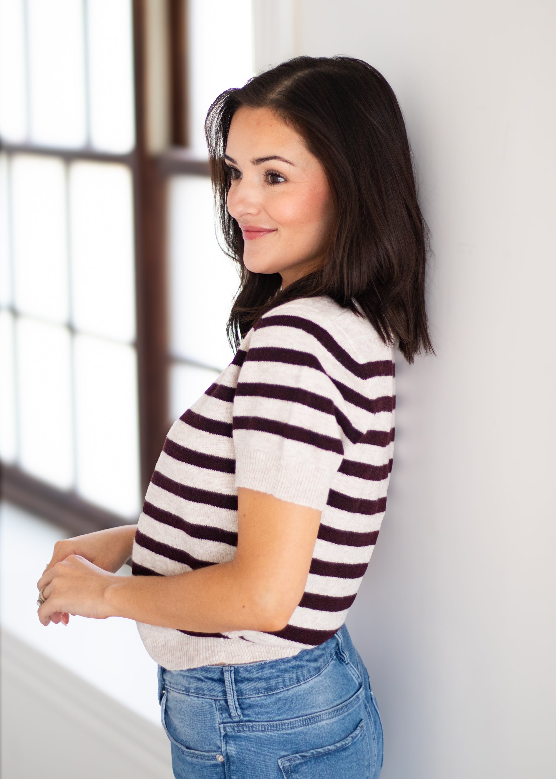 A woman with dark hair leans against a white wall next to a window, wearing a short-sleeve knit top with dark brown and off-white horizontal stripes, paired with blue jeans.