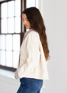 A woman with long brown hair is shown from the side, wearing a cream-colored jacket over a black and white checkered shirt, paired with blue jeans, in a bright room with a large window.