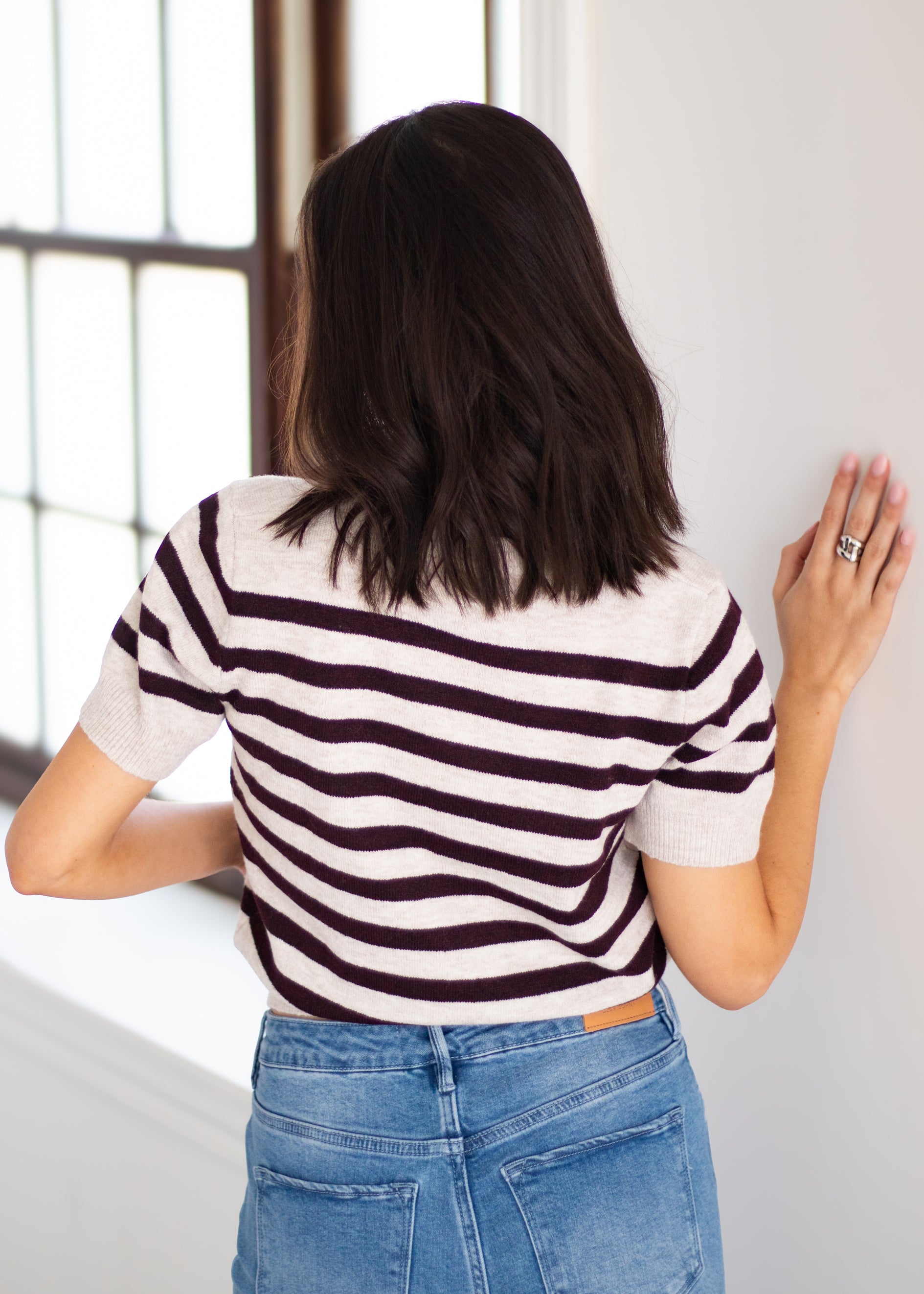 A woman viewed from behind wearing a short-sleeve knit top with dark brown and off-white horizontal stripes, paired with blue jeans.